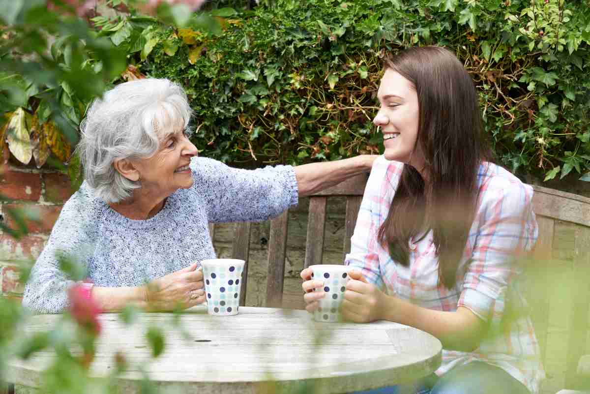 An elderly woman with short gray hair and a joyful smile chats with a younger woman in a plaid shirt as they sit together on a wooden bench in a lush garden, each holding a polka-dotted mug.