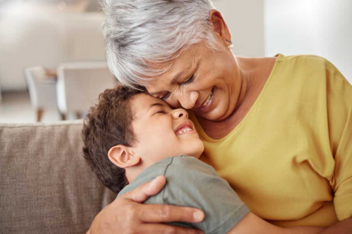 A joyful grandmother with short gray hair wearing a mustard yellow top hugs her young grandson tightly on a couch. Both are smiling with their eyes closed, pressing their foreheads together in a warm, affectionate moment.