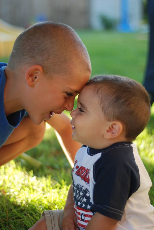 Two young brothers sit on the grass in a backyard, joyfully pressing their foreheads together and smiling. The older boy leans in with a playful expression while the younger toddler looks up at him with a big grin.