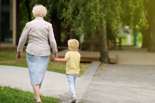 A grandmother and her young grandchild walk hand-in-hand through a park. The grandmother, with short white hair and wearing a light blouse and denim skirt, walks alongside the child in a yellow shirt and light blue pants. Their backs are to the camera as they stroll down a path surrounded by trees.