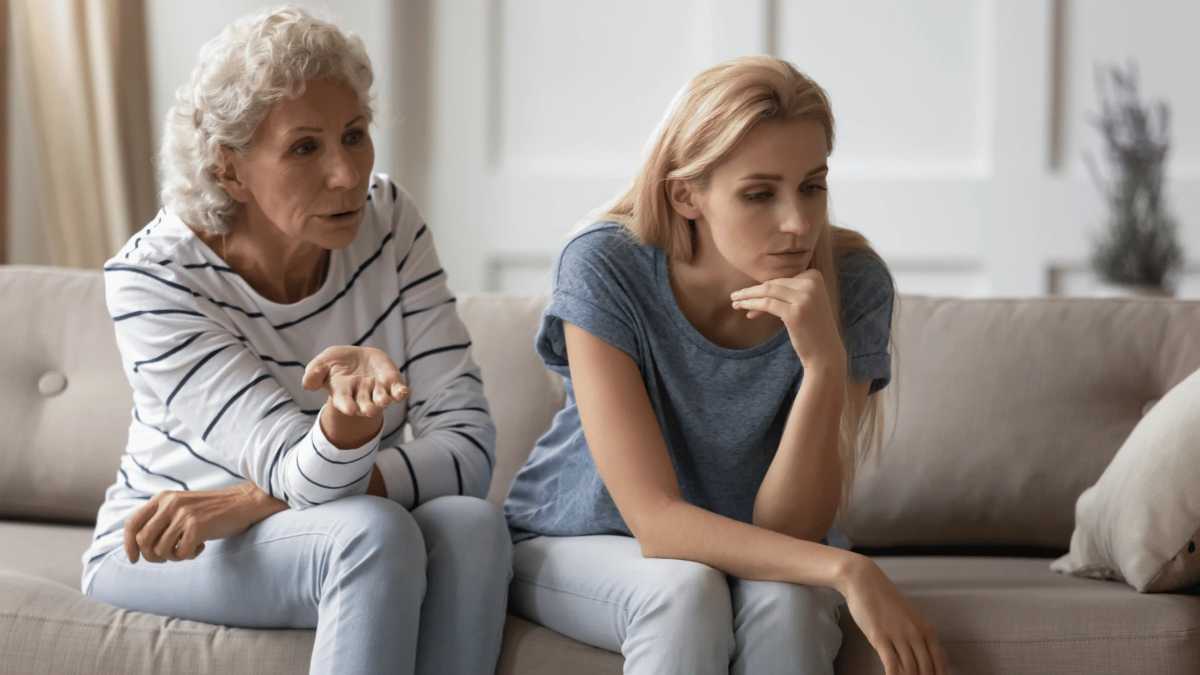 An older woman with curly gray hair is talking to a younger woman with long blonde hair on a couch. The older woman gestures with her hand, appearing concerned or expressive, while the younger woman sits with her chin resting on her hand, looking upset or deep in thought. The mood appears tense or emotional.