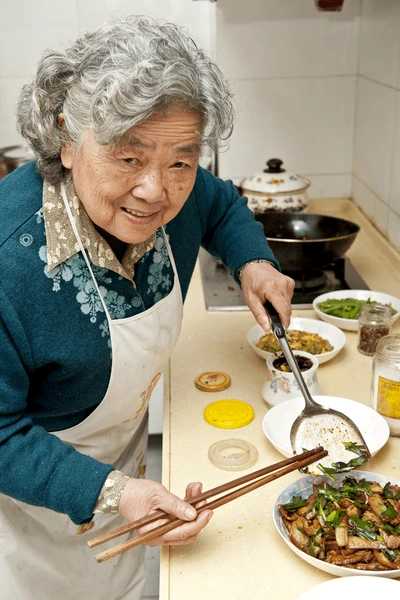 An elderly woman with curly gray hair smiles warmly while preparing a meal in the kitchen, holding chopsticks in one hand and serving rice with the other.