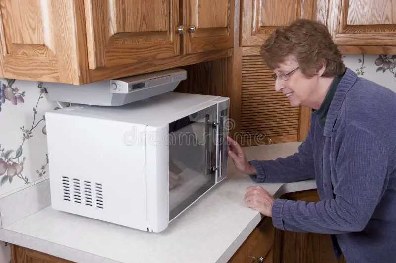 A senior woman in a cozy kitchen leans forward to open a microwave, watching it closely with a gentle smile.
