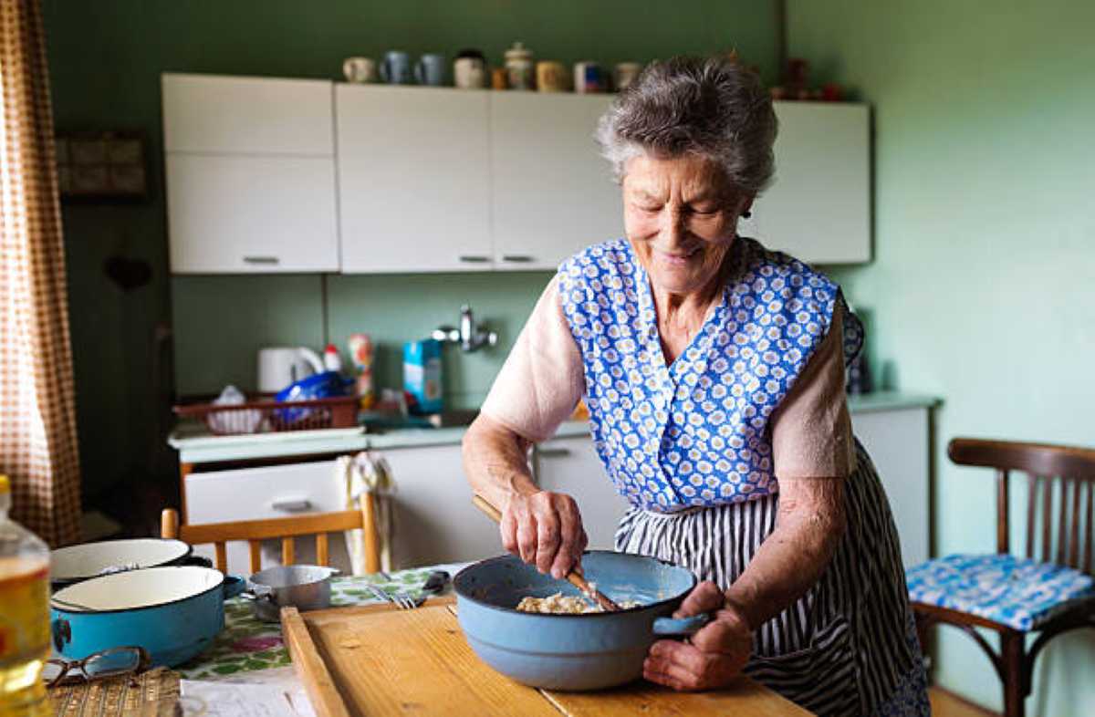 An older woman in a floral apron stirs ingredients in a large blue mixing bowl on a wooden kitchen table, surrounded by vintage cookware.