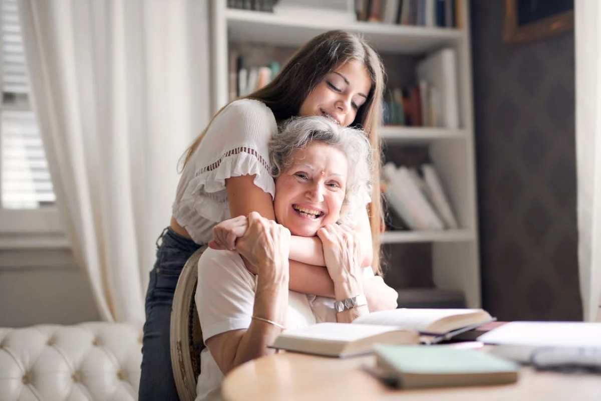 A joyful elderly woman with gray hair sits at a table with open books while her smiling granddaughter lovingly hugs her from behind in a cozy room filled with bookshelves and soft light.
