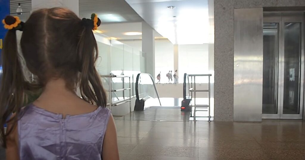 Little girl with pigtails and a purple dress stands alone, looking toward an airport escalator.