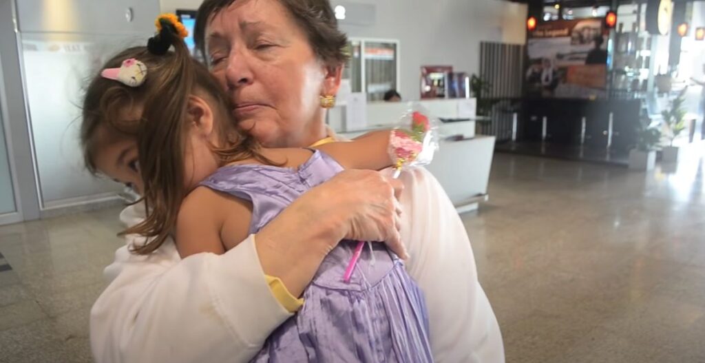 Older woman tearfully hugs a young girl tightly while holding a small wrapped flower at the airport.