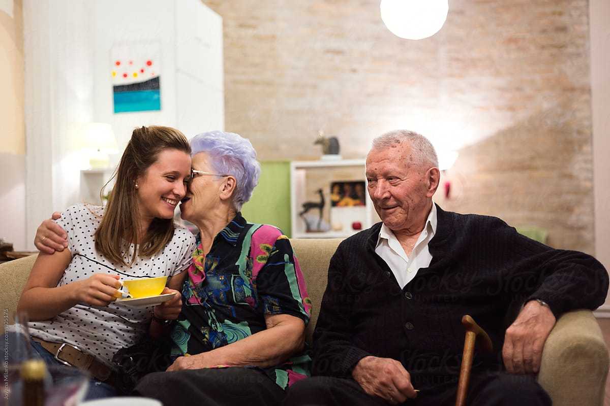 A young woman holding a yellow teacup sits on a beige couch, laughing as an elderly woman with short lavender hair whispers in her ear and hugs her. An elderly man with a cane, wearing a black sweater, sits beside them smiling warmly in a cozy, softly lit living room.