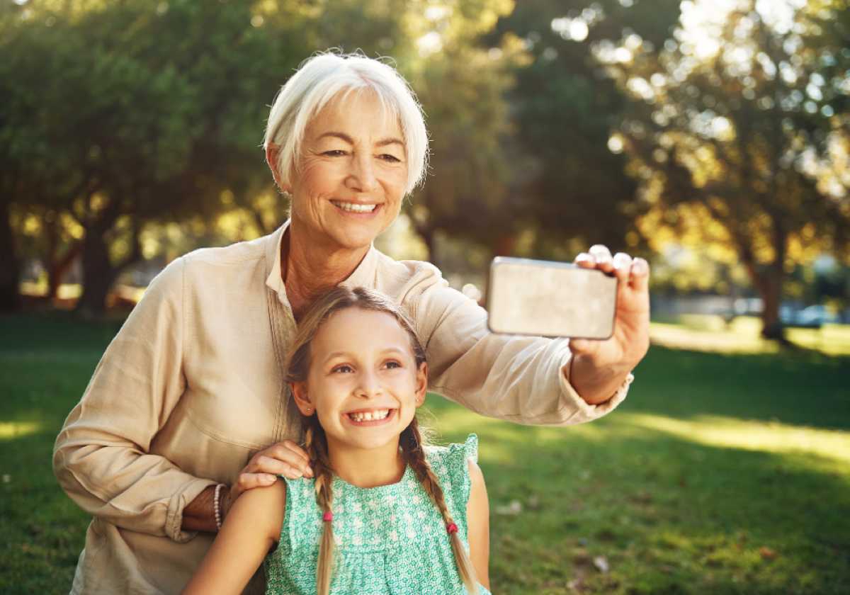 Smiling grandmother takes a selfie with her granddaughter in a sunny park, both looking happy and enjoying time outdoors.