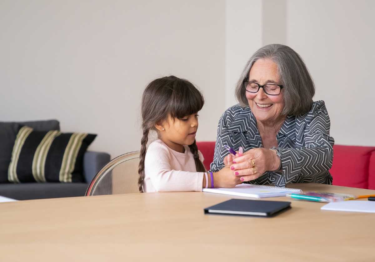 Smiling grandmother helping her young granddaughter write with a pencil at a table, enjoying a creative moment together.