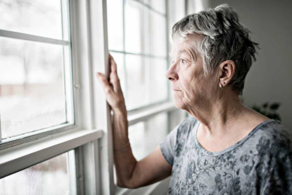 Elderly woman with short gray hair looks out a window with a thoughtful expression, resting one hand on the frame.