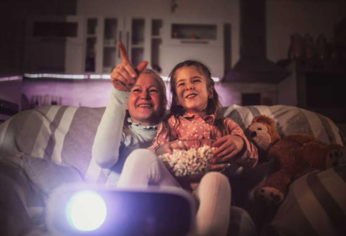 A grandmother and her young granddaughter sit on a striped couch, smiling and pointing at a projected movie while holding a bowl of popcorn, with a teddy bear resting beside them.