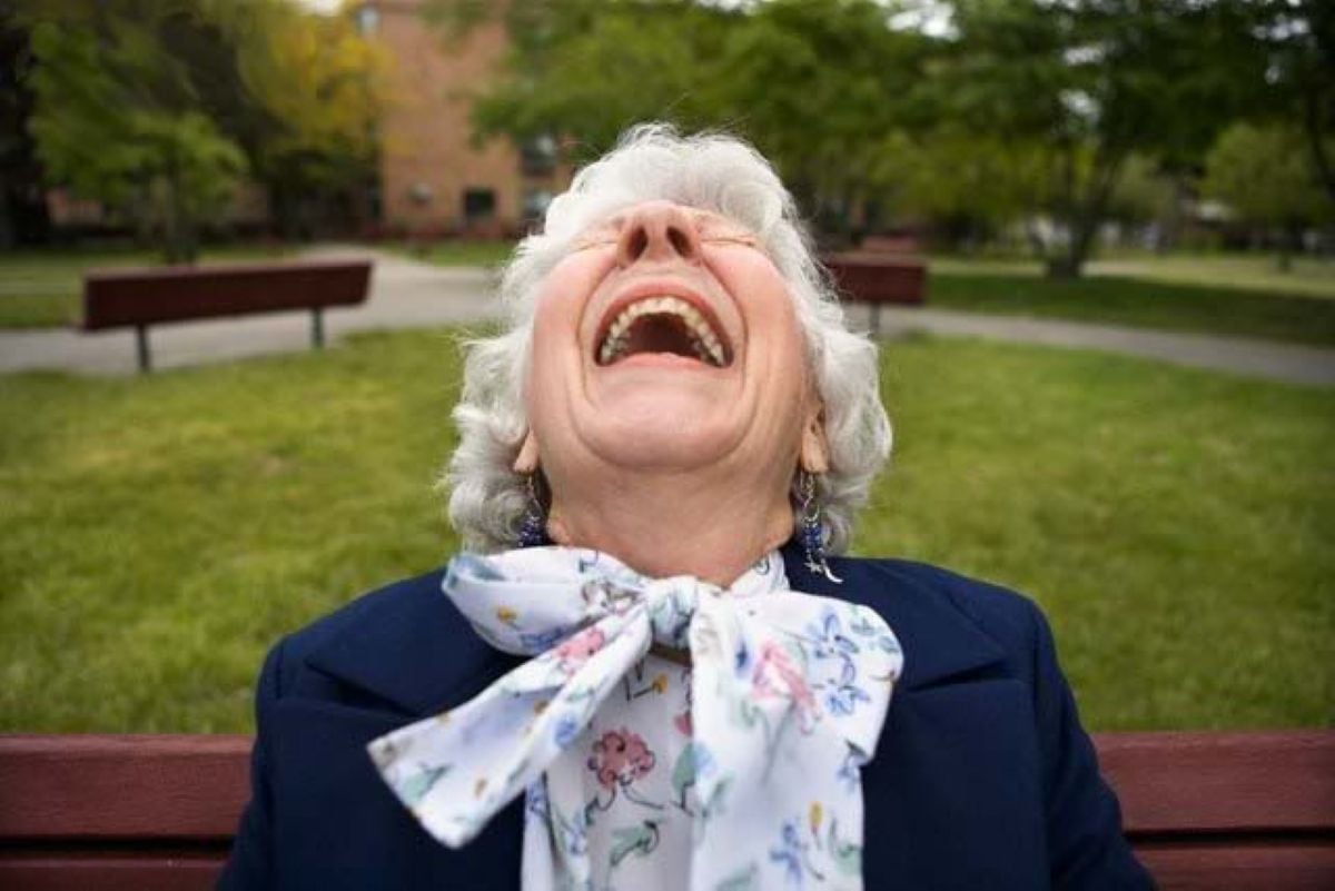Elderly woman sitting on a park bench, leaning back with her head up and laughing joyfully.