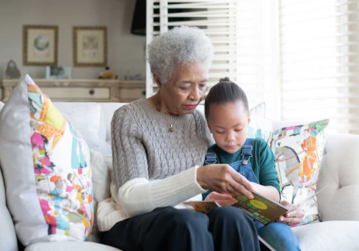 Elderly woman and young girl sit on a couch reading a book together in a bright living room.