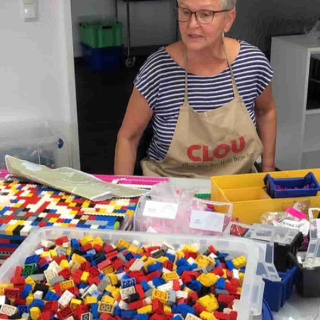 Woman wearing an apron sorts colorful LEGO bricks while assembling accessibility ramps at a crafting table.