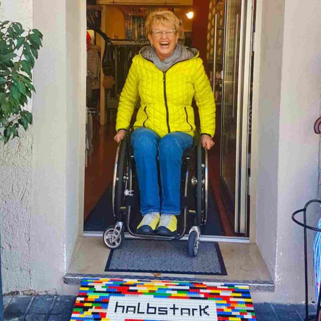 Cheerful woman in a yellow jacket uses a wheelchair to exit a shop via a LEGO brick ramp labeled "halbstark."