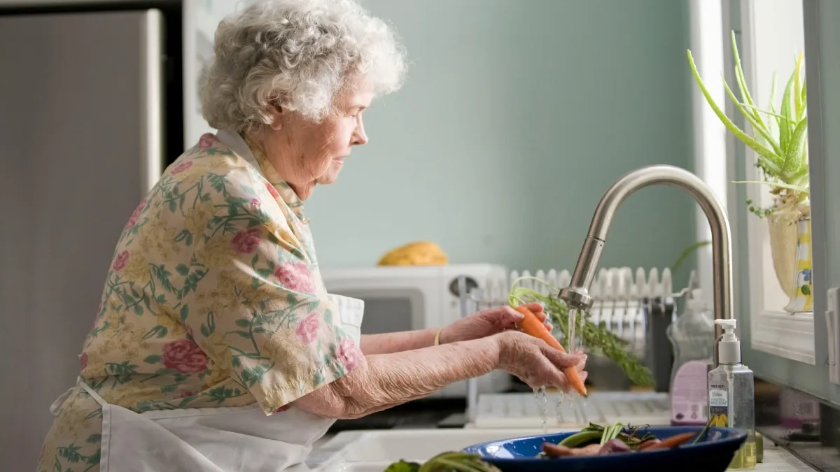An elderly woman washes carrots under a kitchen faucet, with a dish rack and potted plant visible in the background.