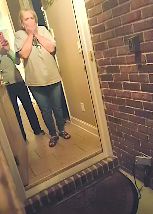 Woman stands shocked and teary-eyed at the front door, hands covering her mouth in surprise.