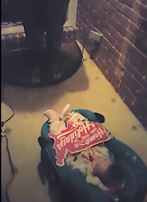 Baby in a carrier on a porch with a festive “Home for the Holidays” sign placed on top.