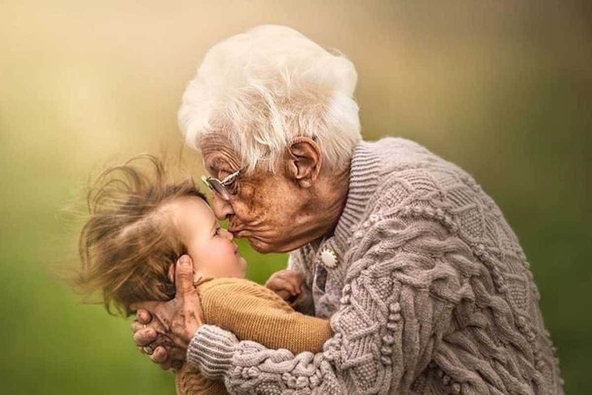 Elderly woman with white hair kisses a young child on the nose while hugging them outdoors.