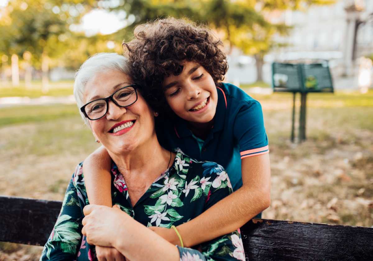 A smiling grandmother with short white hair and glasses sits on a park bench while her curly-haired grandson hugs her from behind, both looking joyful and close in the warm afternoon light.