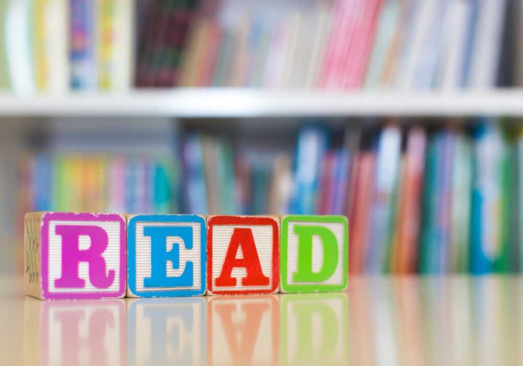 Colorful letter blocks spelling "READ" sit on a reflective surface with a blurred bookshelf in the background.