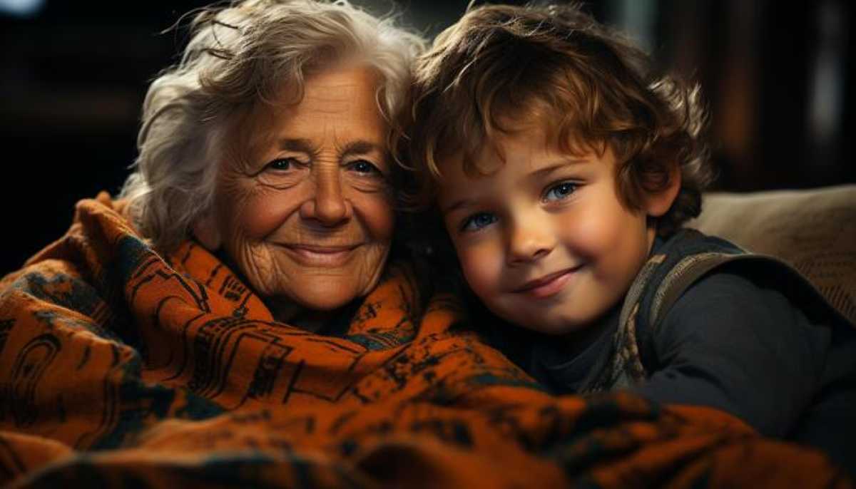Smiling grandmother and young boy cuddling under a cozy orange blanket, sharing a warm moment indoors.