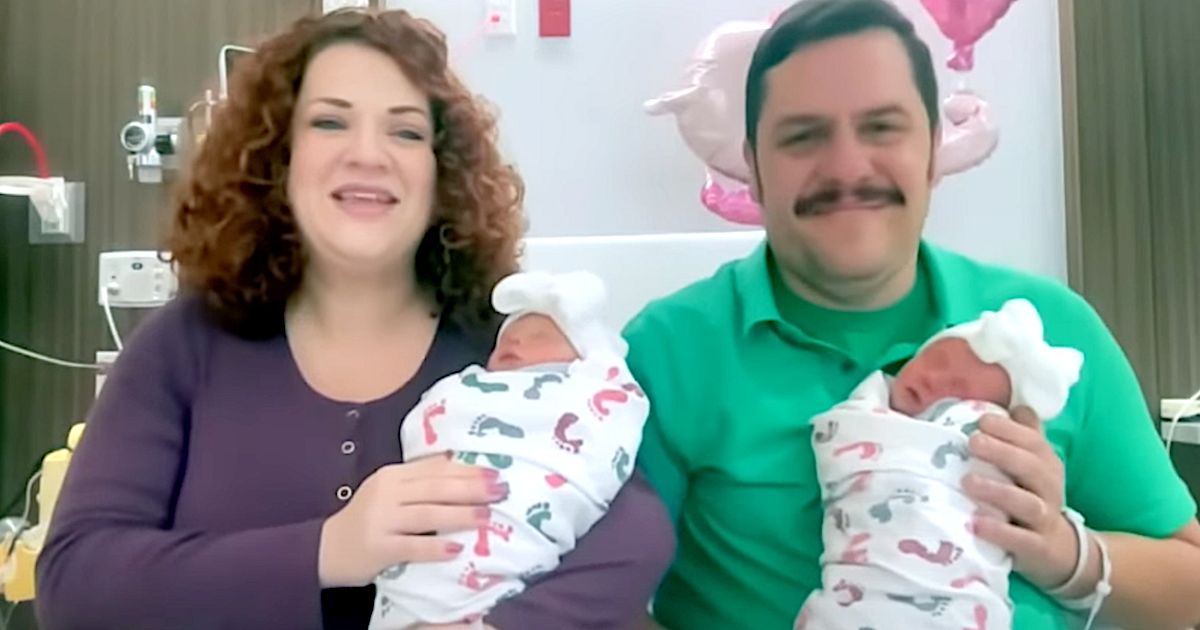 Smiling parents sit in a hospital room holding newborn twins wrapped in patterned blankets with white bows on their heads.
