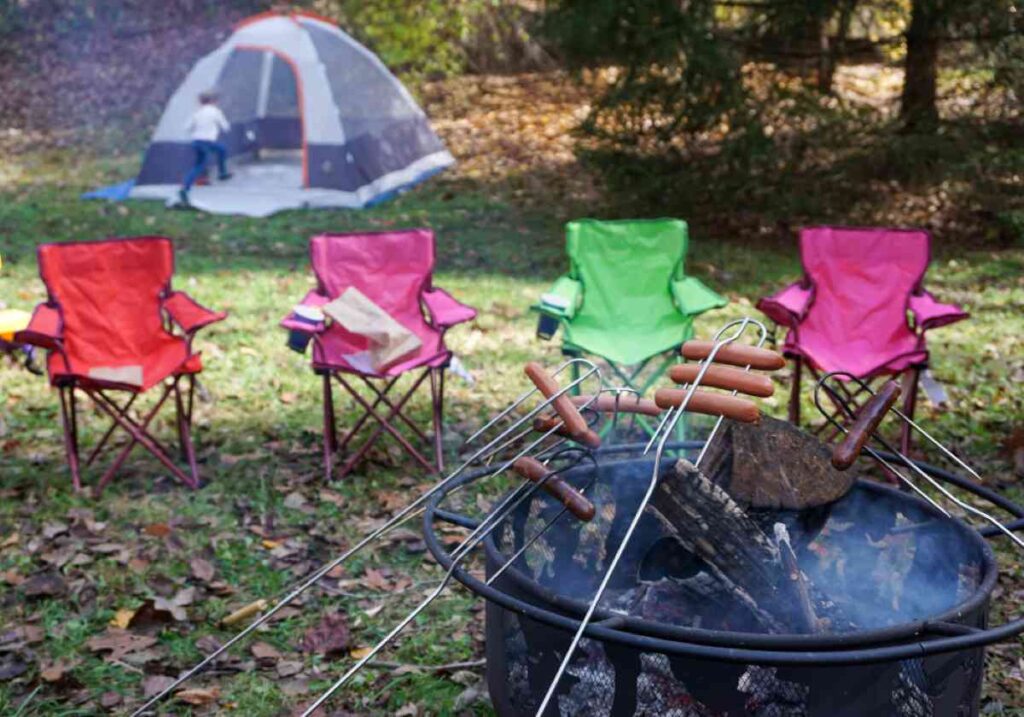 Camping scene with hot dogs roasting over a firepit, colorful chairs arranged in a circle, and a child running toward a tent in the background.