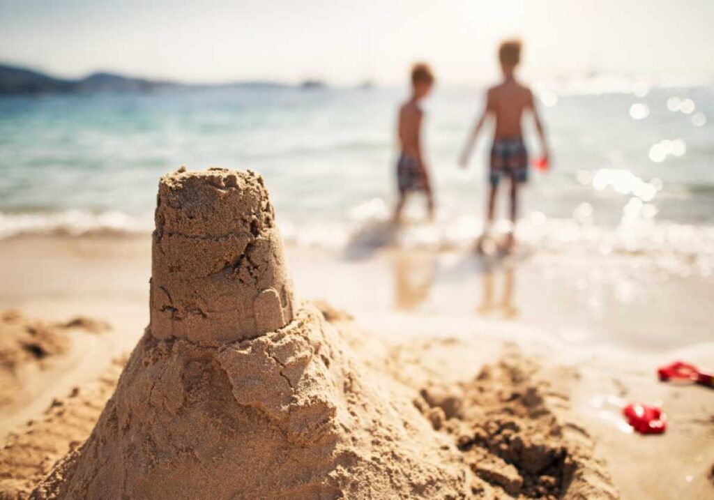 Close-up of a sandcastle on the beach with two children playing in the ocean in the blurred background.