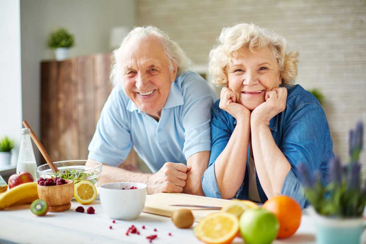 Smiling senior couple in the kitchen surrounded by fresh fruits, enjoying a healthy moment together.