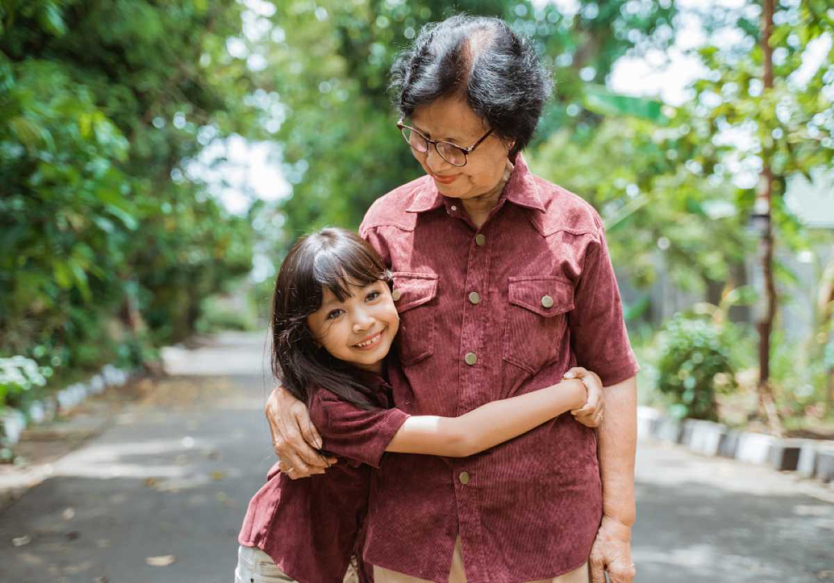 A happy young girl hugs her smiling grandmother while standing on a tree-lined path, both wearing matching maroon shirts.