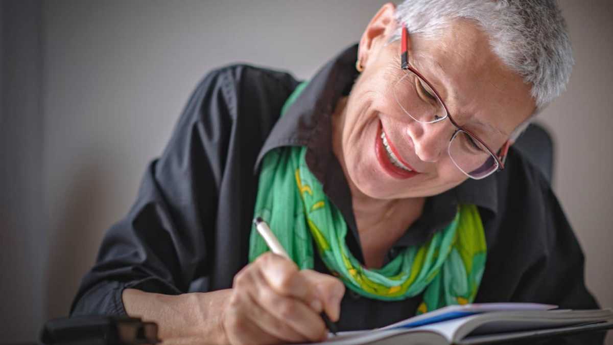 A smiling older woman with short gray hair and glasses writes in a notebook while sitting at a table, wearing a black shirt and a bright green scarf.