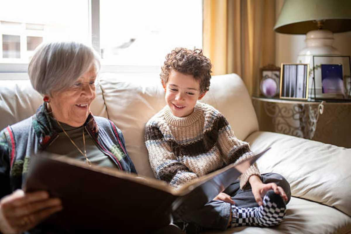Smiling grandmother and young boy sit on a couch looking through a photo album together in a cozy living room.