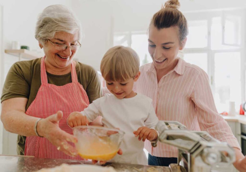 Smiling grandmother, mother, and young boy mix eggs in a glass bowl together in a bright kitchen.