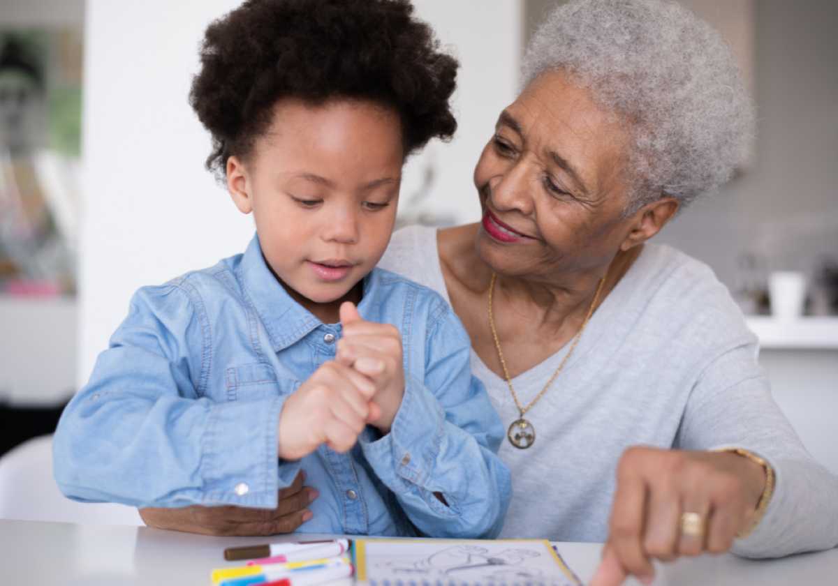 Elderly woman watches a young boy color in a book at a table, smiling warmly.