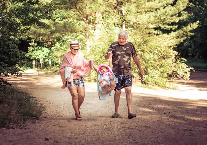 Grandparents joyfully swing a toddler between them while walking on a sunny forest path.