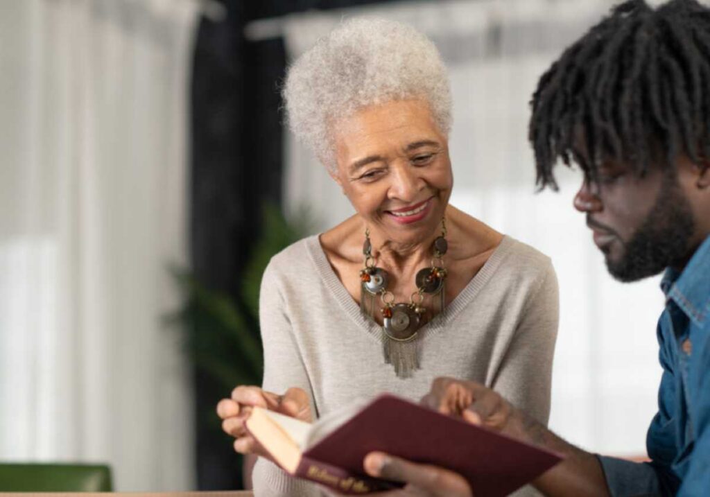 Elderly woman and young man share a book, smiling as they read together at a table.