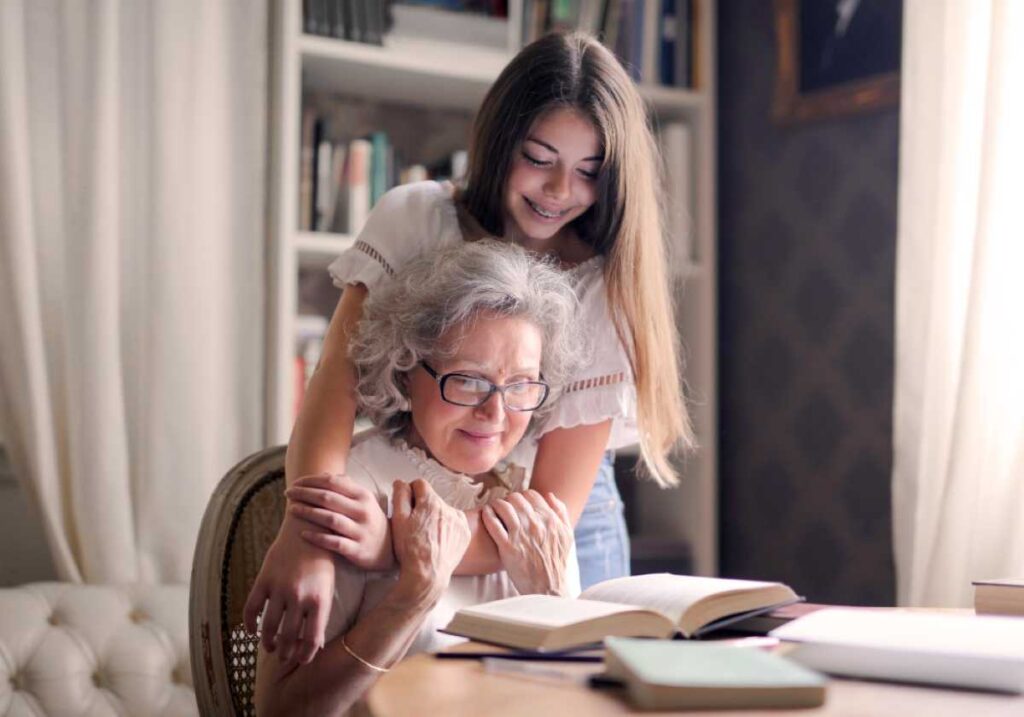 Teen girl hugs her grandmother from behind while reading a book in a cozy library.