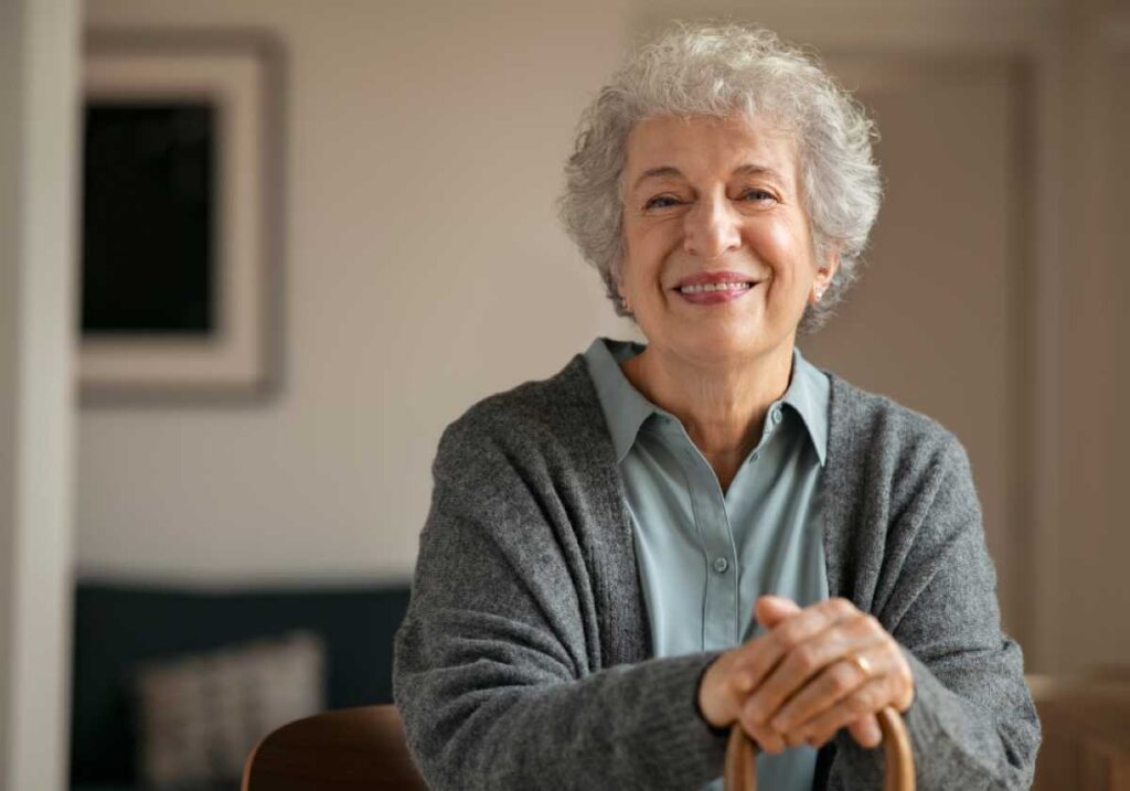 Smiling elderly woman with gray curly hair in a gray cardigan sits indoors, hands resting on a cane.