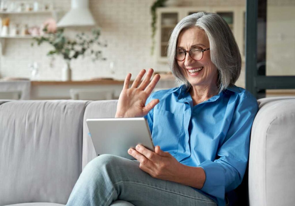 Smiling elderly woman with glasses waves while holding a tablet on a couch at home.