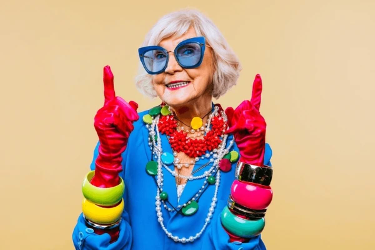 A stylish older woman wearing oversized blue sunglasses, bright red gloves, chunky colorful jewelry, and a vibrant blue outfit, smiling confidently against a light yellow background.
