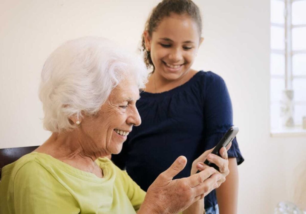 The Cool Grandma’s Guide to Trends: Connecting With Your Grandkids in Today’s World 2 A happy grandmother holding a smartphone while her smiling granddaughter stands beside her, helping her use the device.