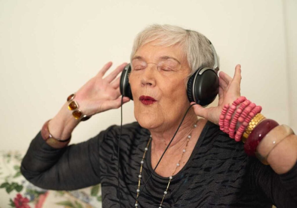 An older woman wearing large headphones, eyes closed, and smiling while enjoying music, accessorized with colorful bracelets and a black top.