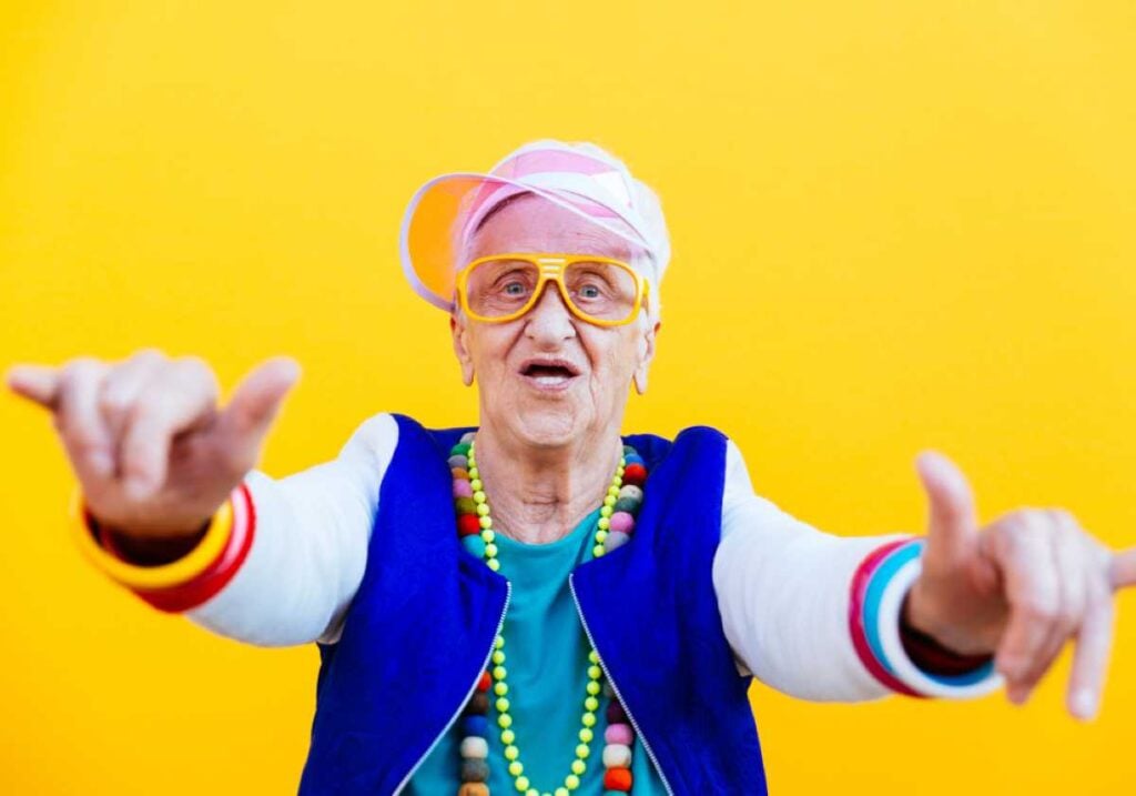 A cheerful older woman dressed in a playful, colorful outfit with a pink visor, yellow glasses, beaded necklaces, and bright bracelets, posing energetically in front of a bold yellow background.