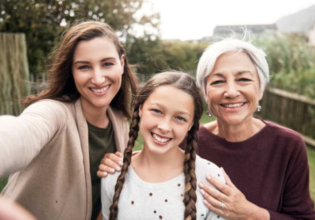 A grandmother, mother, and young granddaughter smiling together outdoors while taking a selfie, showing three generations in one joyful moment.