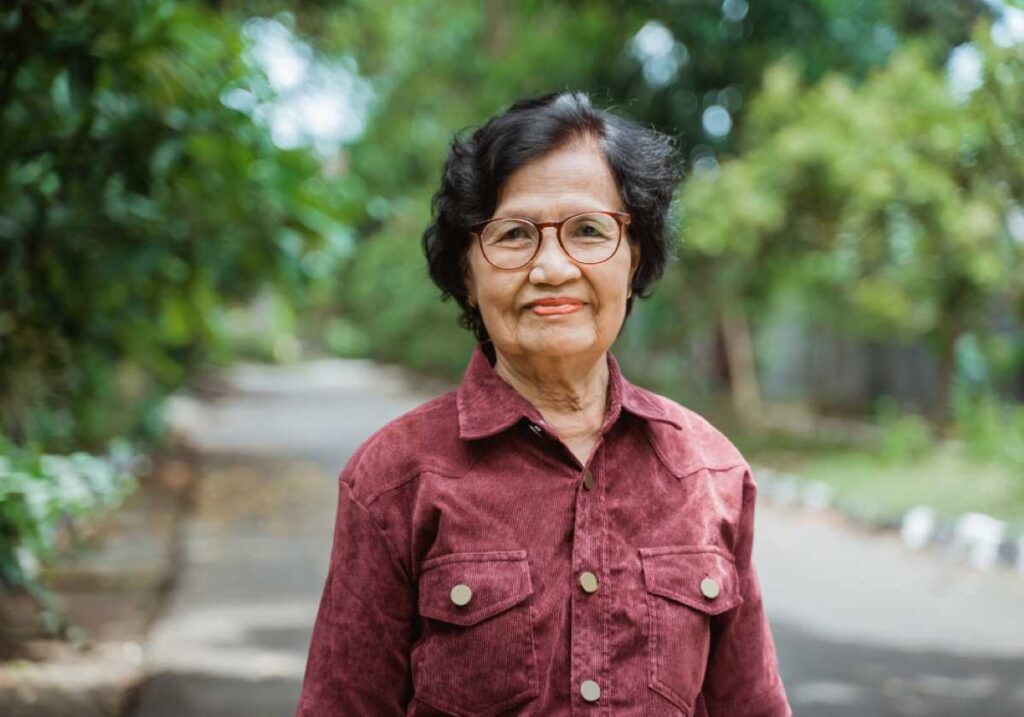 Elderly woman in glasses and a maroon shirt stands outdoors on a sunny day with green trees in the background.