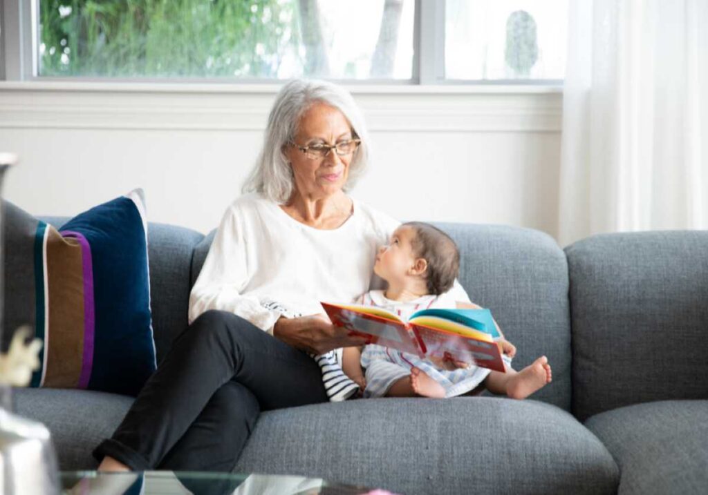 Elderly woman with gray hair reads a colorful book to a baby on a gray couch in a cozy, sunlit living room.