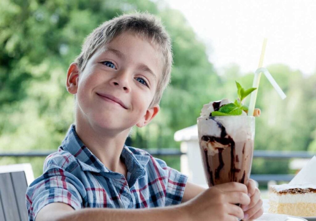 15 Things the Best Grandmas Never Do When Visiting Your Grandkids 2 Smiling boy holding a tall chocolate milkshake topped with whipped cream, mint, and a straw at an outdoor café.