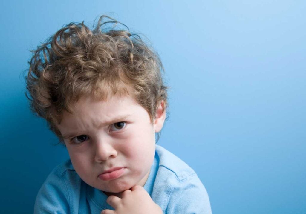 Young boy with curly hair and a pouty face, looking upset or angry while standing against a blue background.